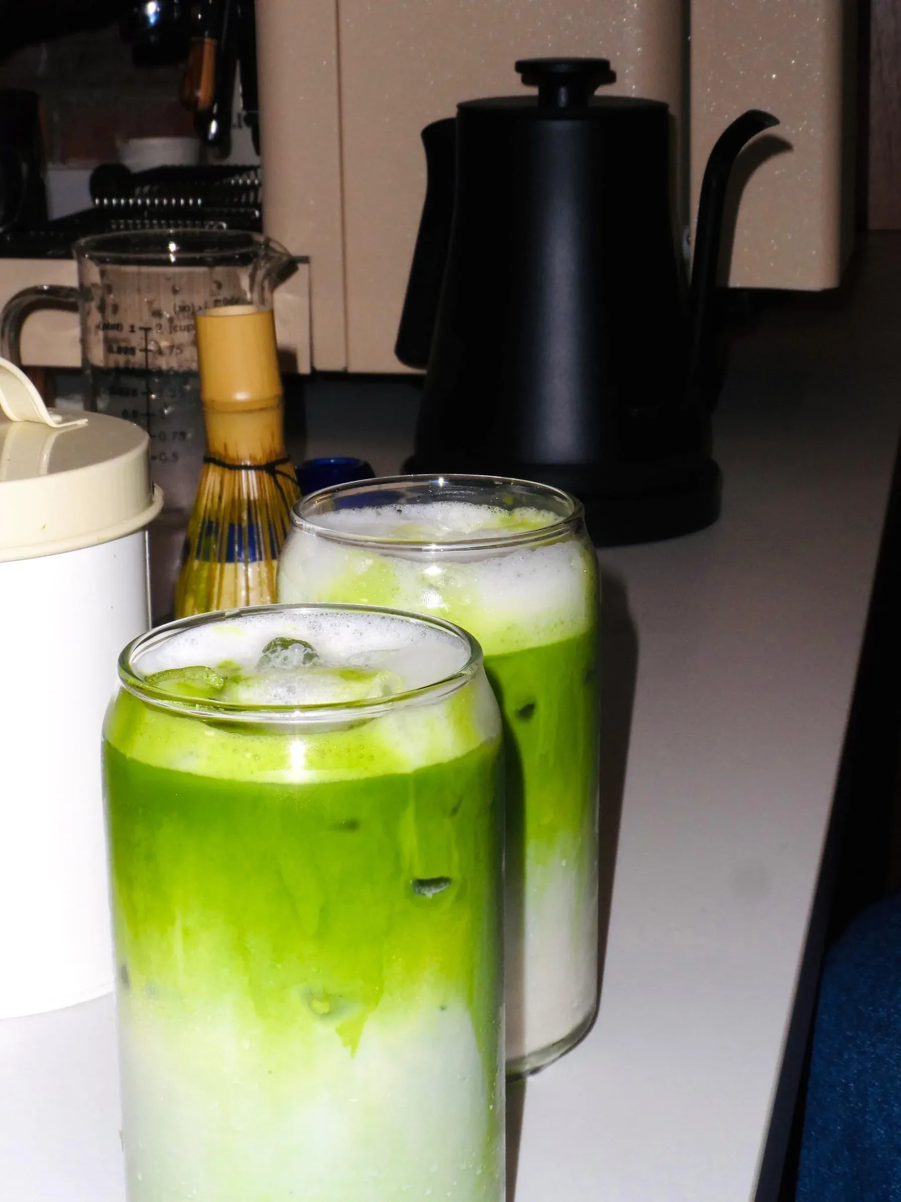 Dos vasos con matcha y leche sobre una mesa, con un hervidor y un chasen al fondo